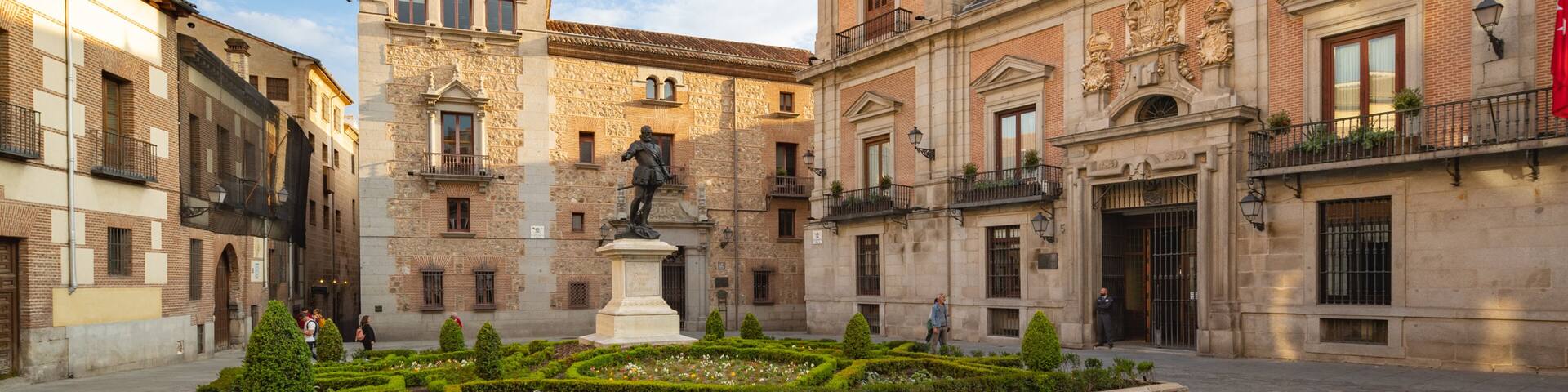 Un patio histórico con una estatua, una iglesia con un campanario y jardines bien cuidados.