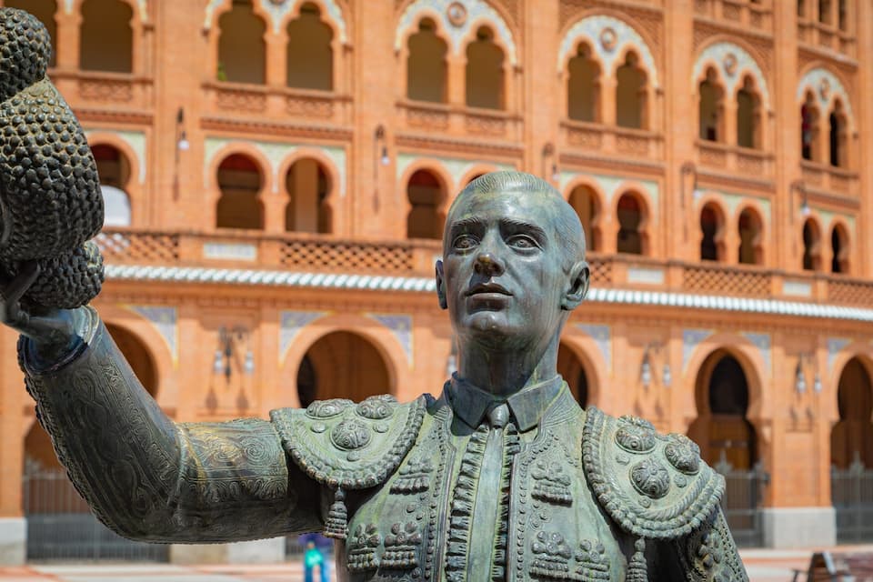 A statue of a man holding a bird in front of a large, ornate building with arches.