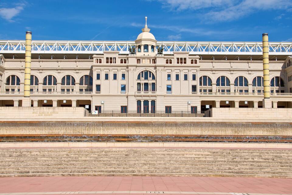 Un grand bâtiment historique doté d’un dôme central et de fenêtres en arc de cercle, un pont avec une structure en treillis au-dessus, et une voie ferrée au premier plan.