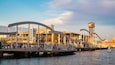A waterfront promenade with a modern bridge, a restaurant, and a tower.