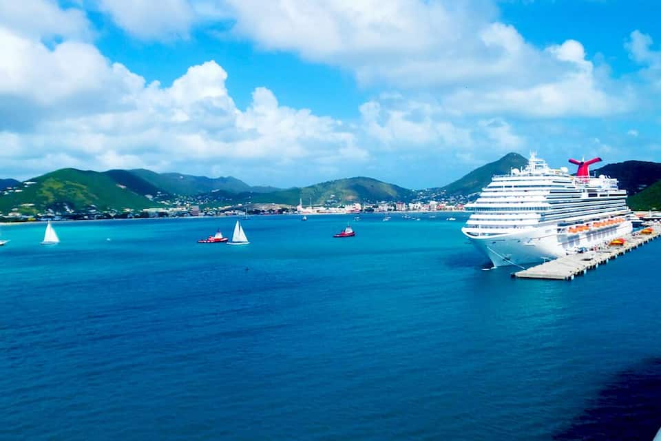Ein großes Kreuzfahrtschiff liegt an einem Steg vor einer Küstenlandschaft mit Bergen und Segelbooten im Hintergrund.