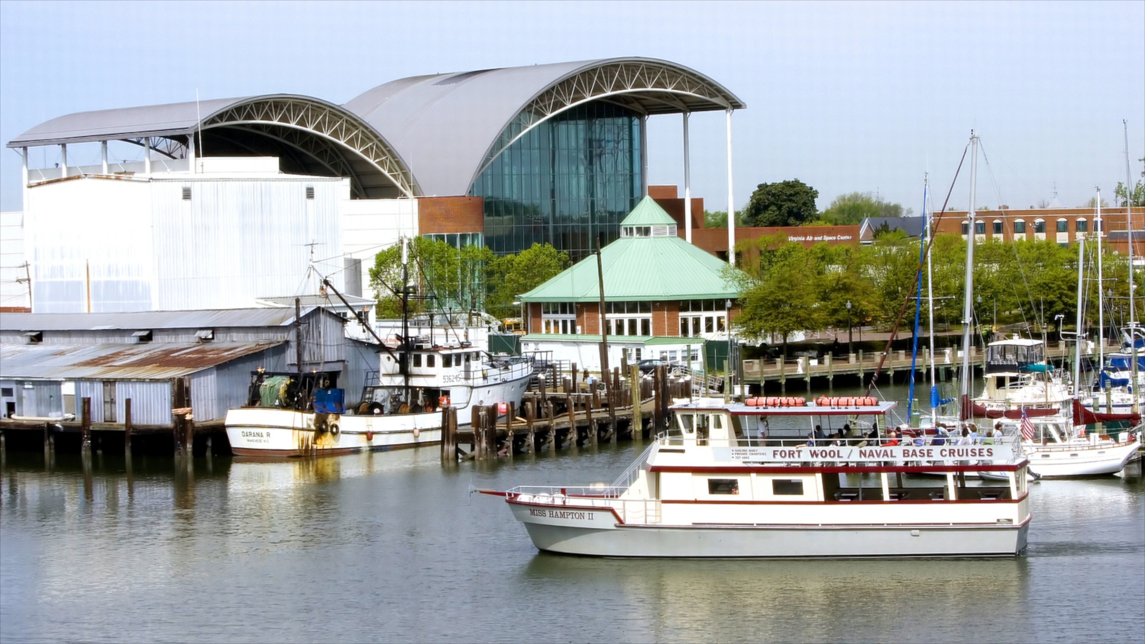 A marina with boats docked, a large modern building with a green roof, and a historic ship named 'DARANA R' in the background.