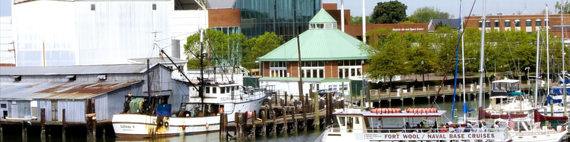 A marina with boats docked, a large modern building with a green roof, and a historic ship named 'DARANA R' in the background.