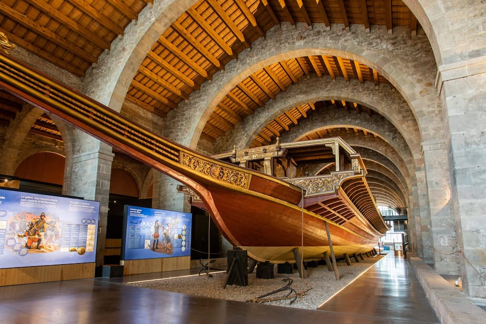 Un grand navire en bois est exposé dans un musée, avec des arches en pierre et des poutres en bois au plafond.