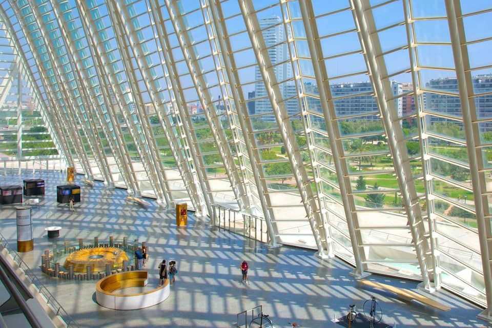 Uma estrutura moderna de vidro com telhado curvo, rodeada por vegetação e edifícios urbanos.