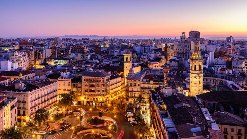 A cityscape at dusk with illuminated buildings, a prominent clock tower, and a roundabout.