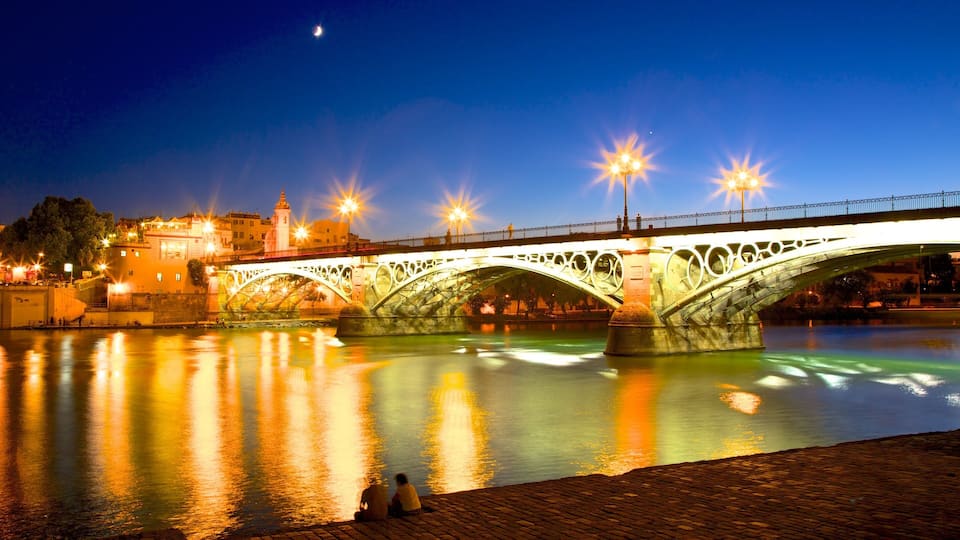 A lit bridge over a calm river at night with a moon in the sky.