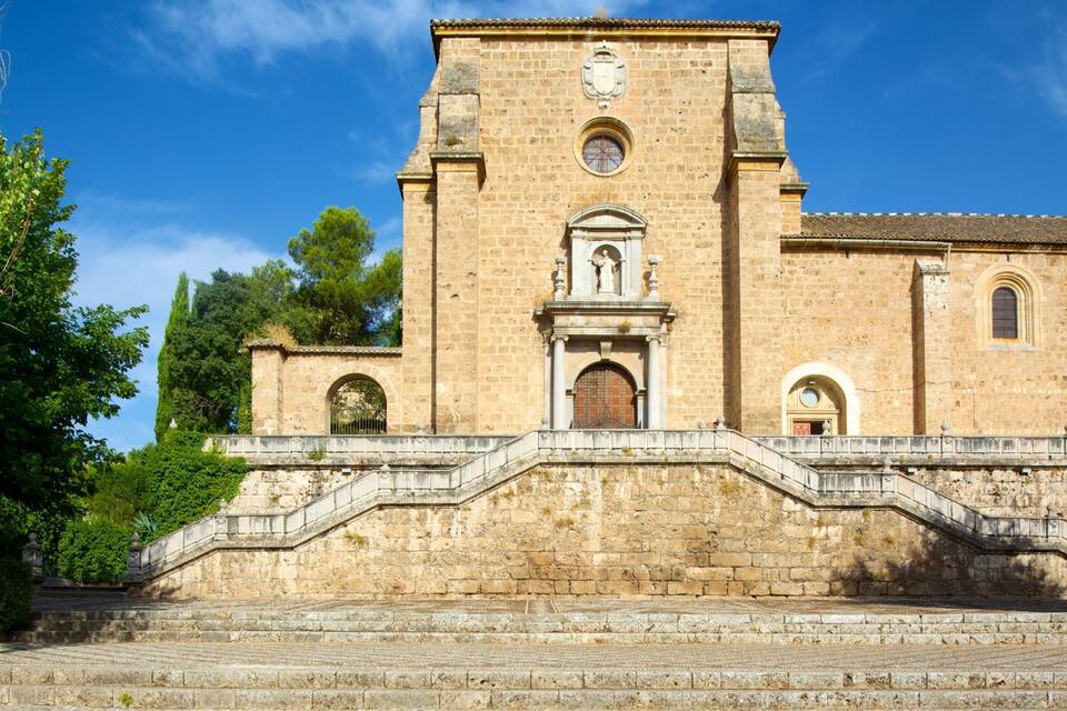 Eine historische Steinkirche mit einem zentralen Glockenturm, einer Treppe, die zum Eingang führt, und üppiger Bepflanzung ringsum.
