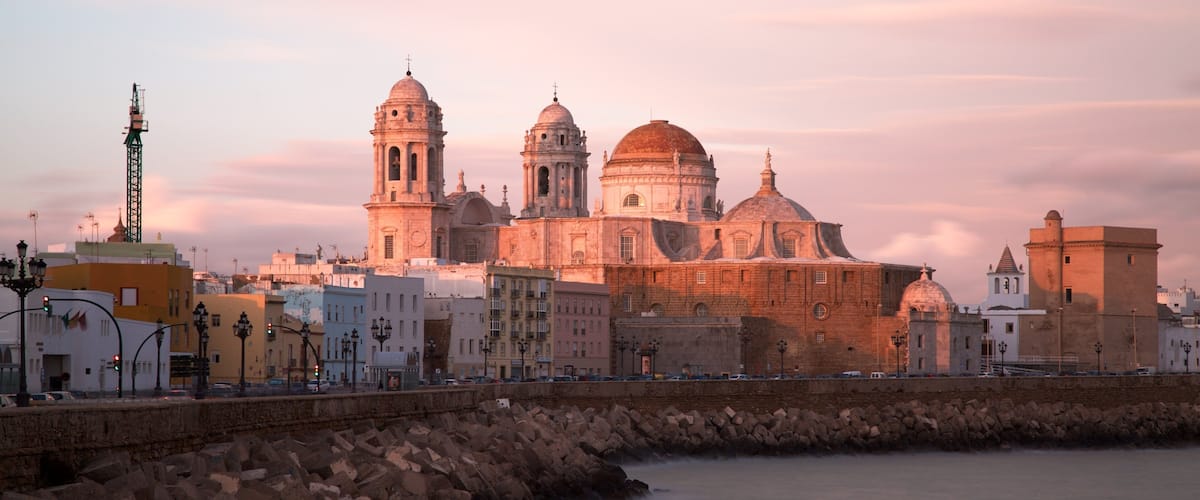 A coastal cityscape with historic architecture, including a prominent church with domes and bell towers, a rocky breakwater, and a calm sea.