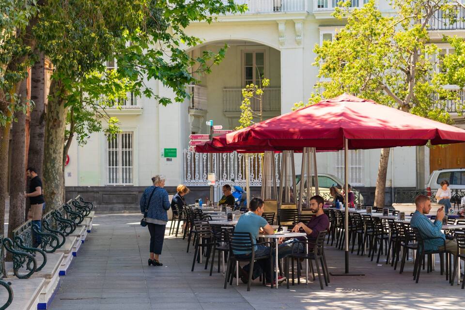 Outdoor cafe seating with red umbrellas, trees, and a building in the background.