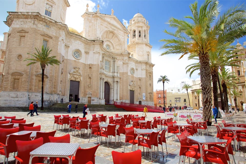 A historic cathedral with two bell towers, palm trees, and red outdoor seating.