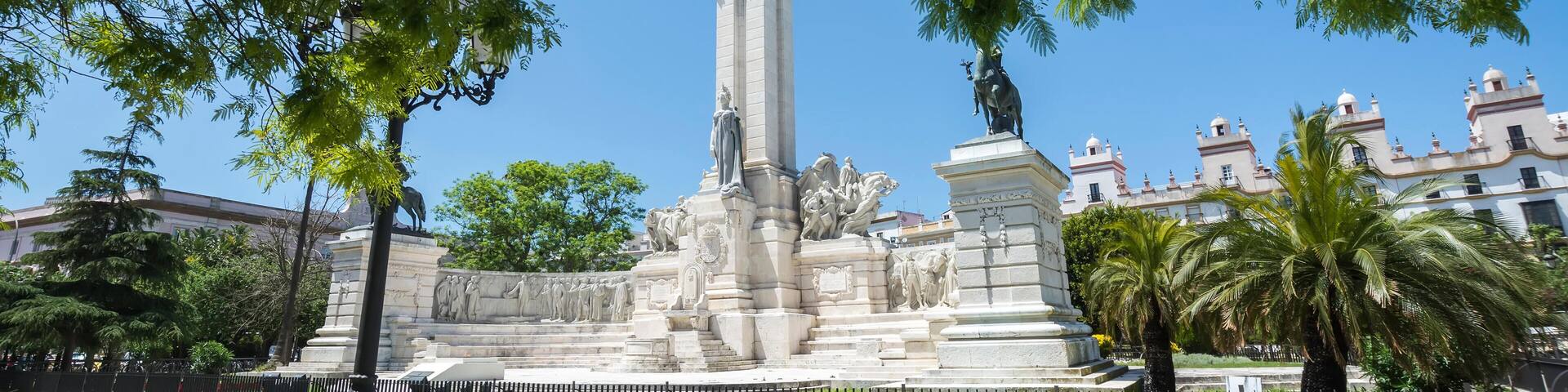 Un monument composé d’une haute colonne et de statues, entouré de palmiers et bordé d’un sol pavé à motifs géométriques.