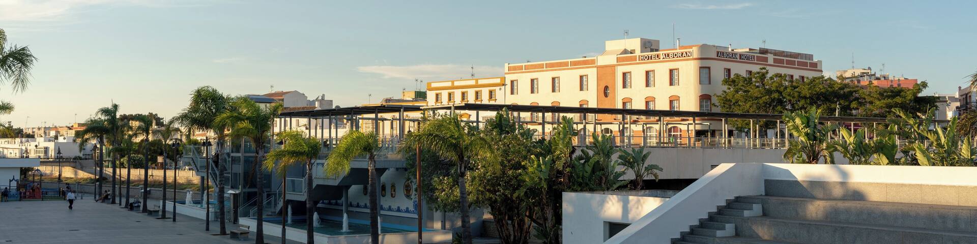 A waterfront promenade with palm trees, a staircase, and a building with a sign.