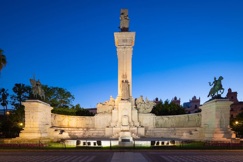 A large monument with a central column and statues on either side, surrounded by a fence and palm trees.