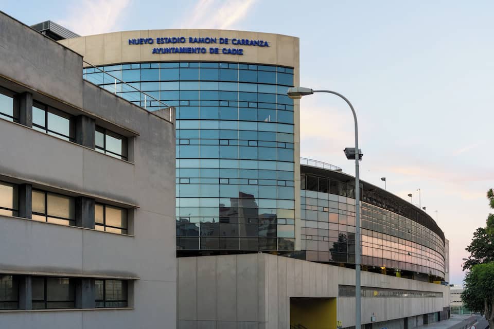 A modern building with a curved glass facade and the text "Nuevo Estadio Ramon de Carranza Ayuntamiento de Cádiz" on the top.