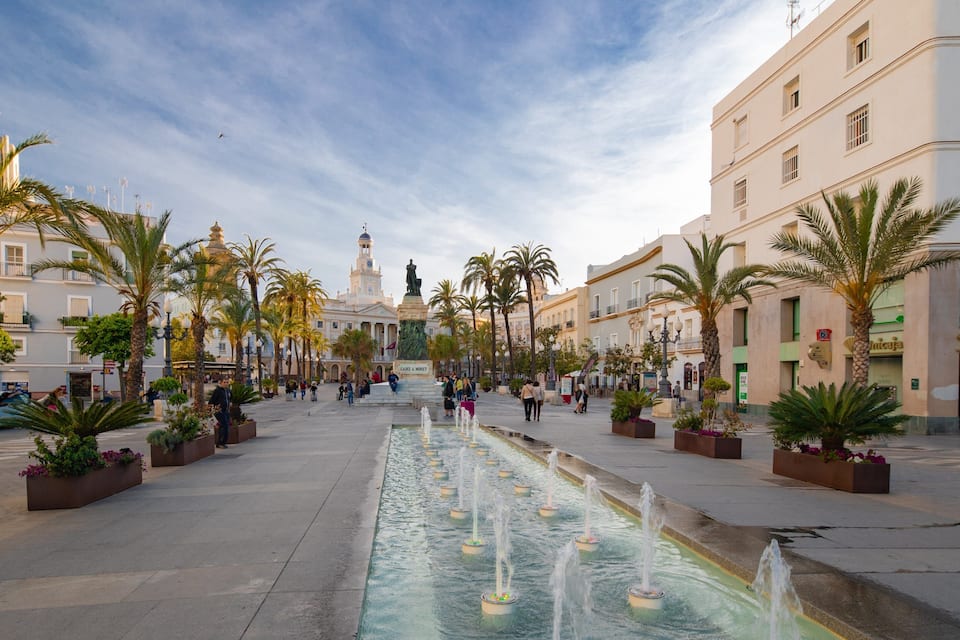 A plaza with a fountain, palm trees, and historic buildings.