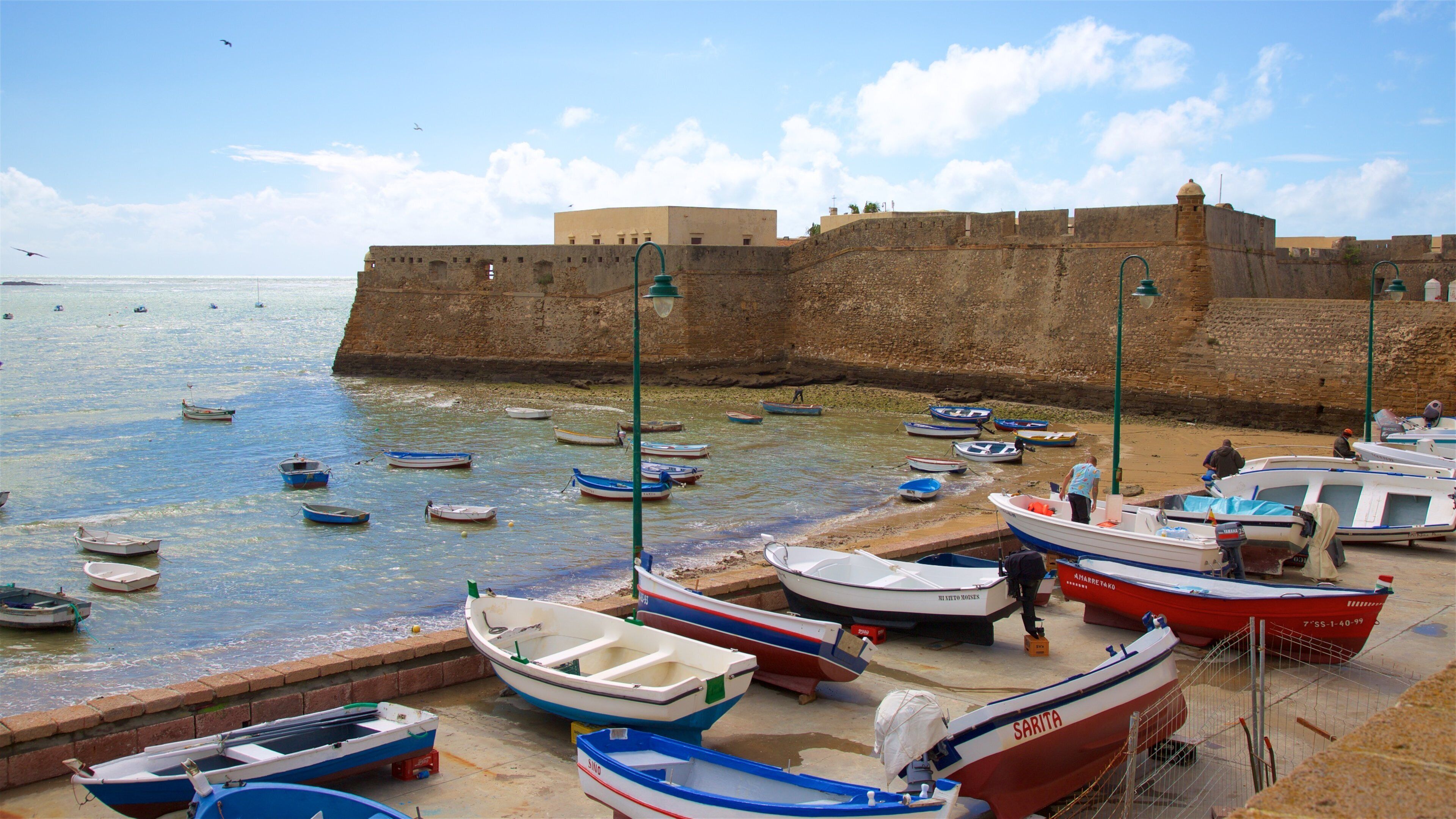 Une scène côtière avec de nombreux petits bateaux amarrés près d’un mur fortifié, au bord de la mer.