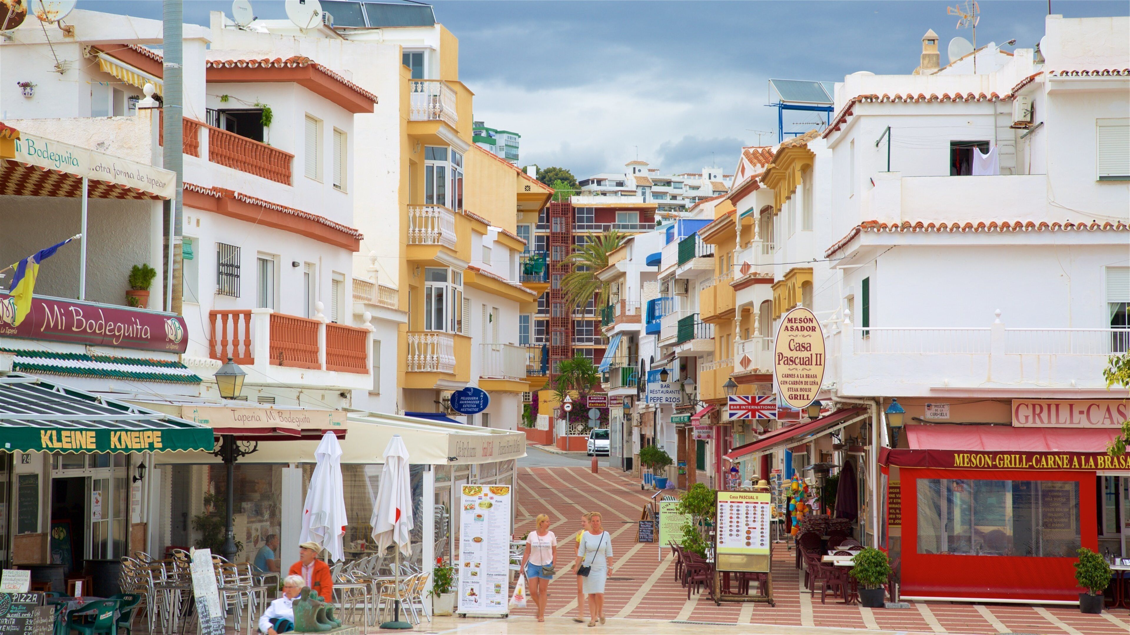 A narrow street lined with colorful buildings, outdoor seating, and various shops and restaurants.