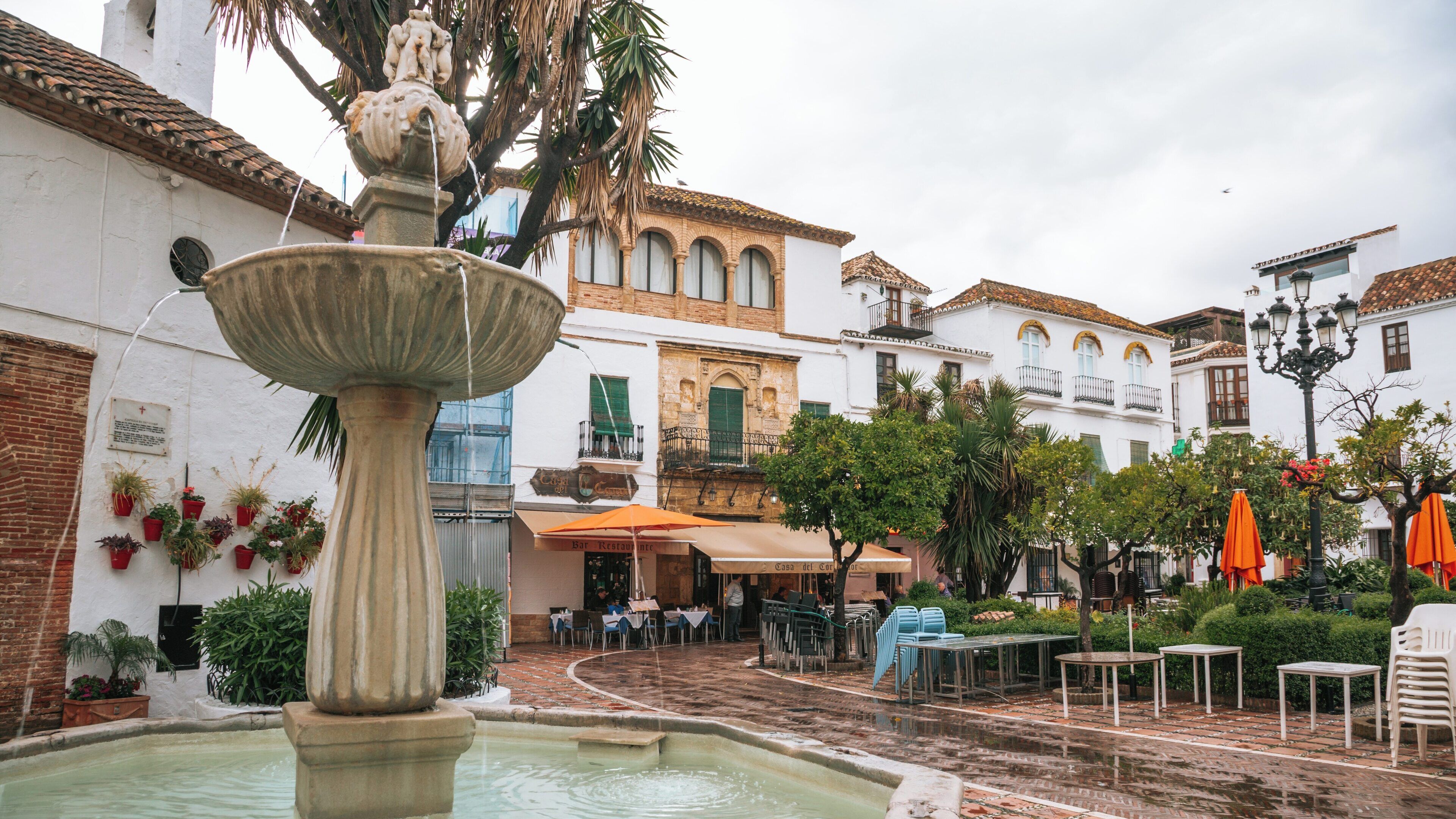 Charming moments in Orange Square, Marbella, a picturesque plaza in Andalusia, Spain, showcasing a fountain, vibrant cafes, and lush greenery under a cloudy sky