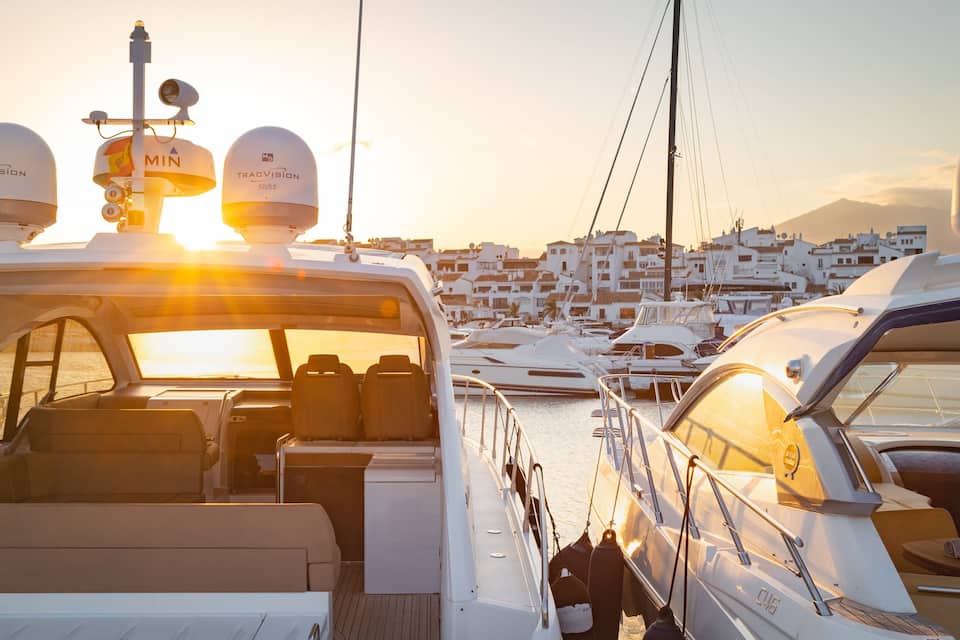 A marina with yachts docked, a sunset in the background, and a cityscape with white buildings.