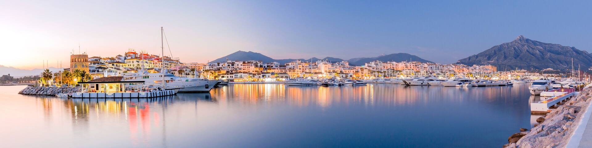 A coastal marina with yachts, a mountain in the background, and a town with colorful buildings.