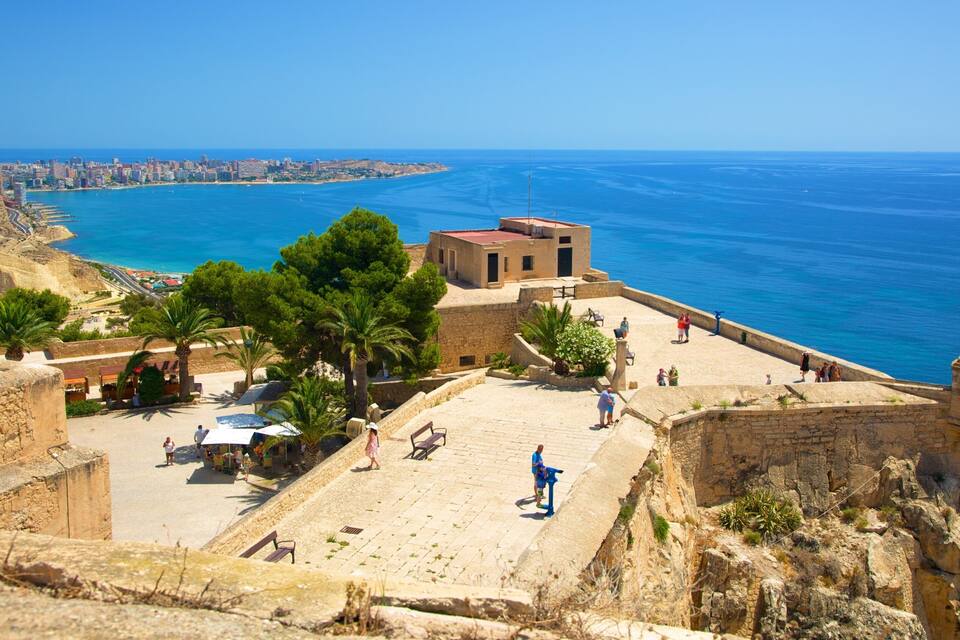 Un paysage urbain côtier avec un fort historique dominant la mer, des palmiers et un ciel bleu dégagé.