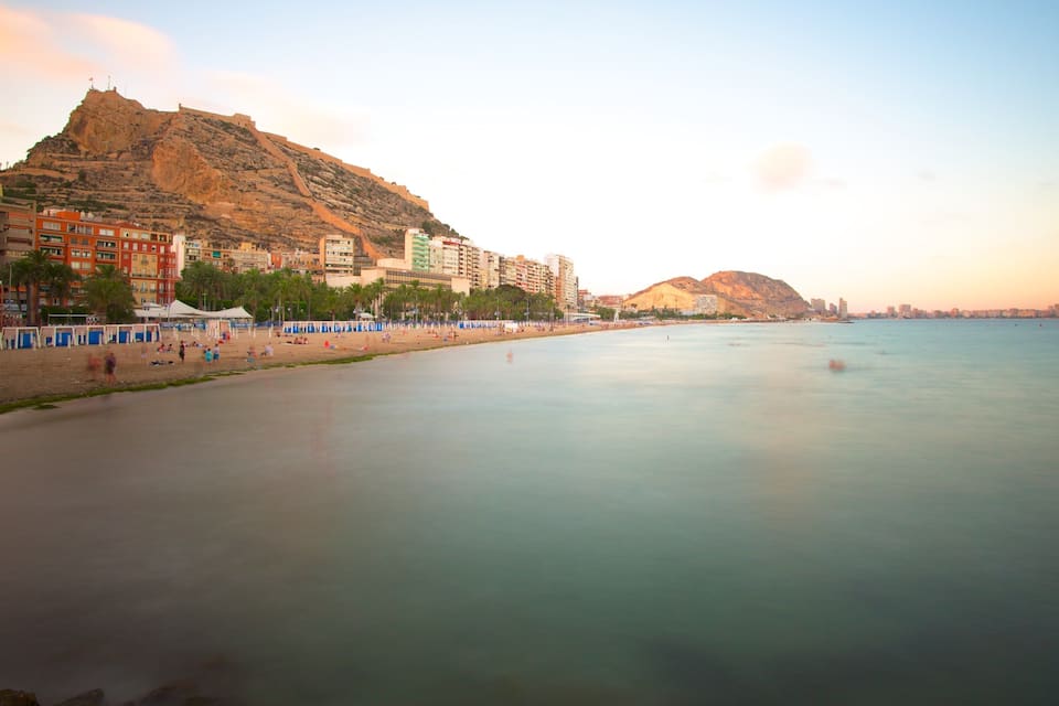 A coastal cityscape with a prominent hill, beach, and calm sea.