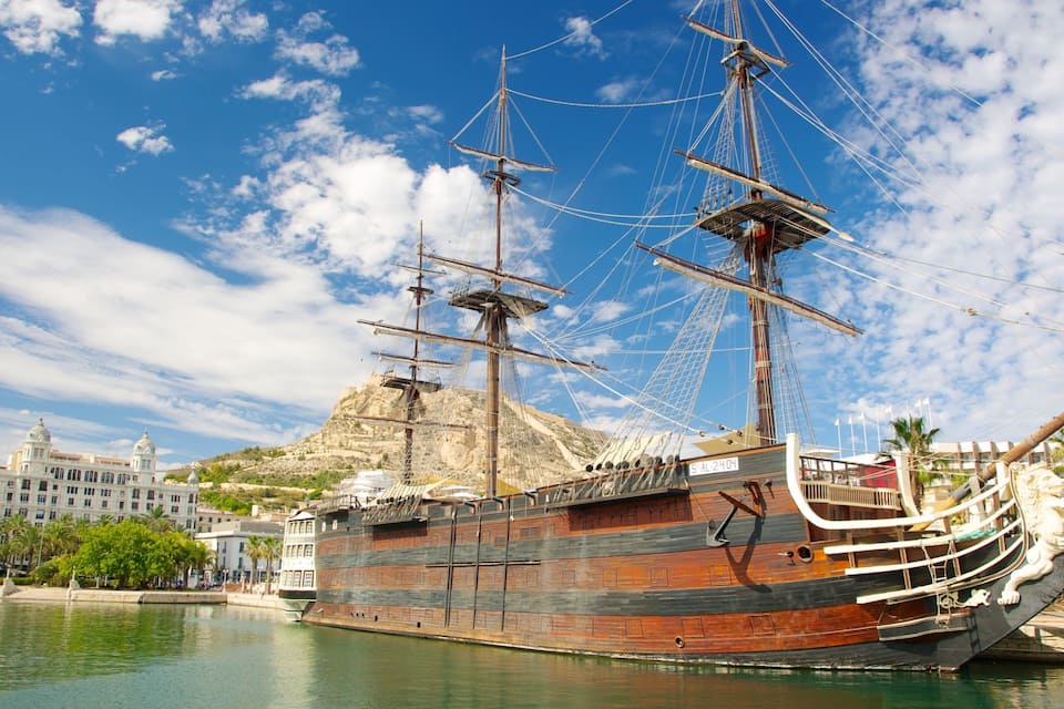 Um grande navio à vela histórico ancorado em um porto, com montanhas ao fundo e edifícios modernos.