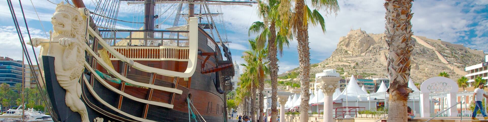 A historic sailing ship docked at a waterfront promenade with palm trees and modern buildings in the background.