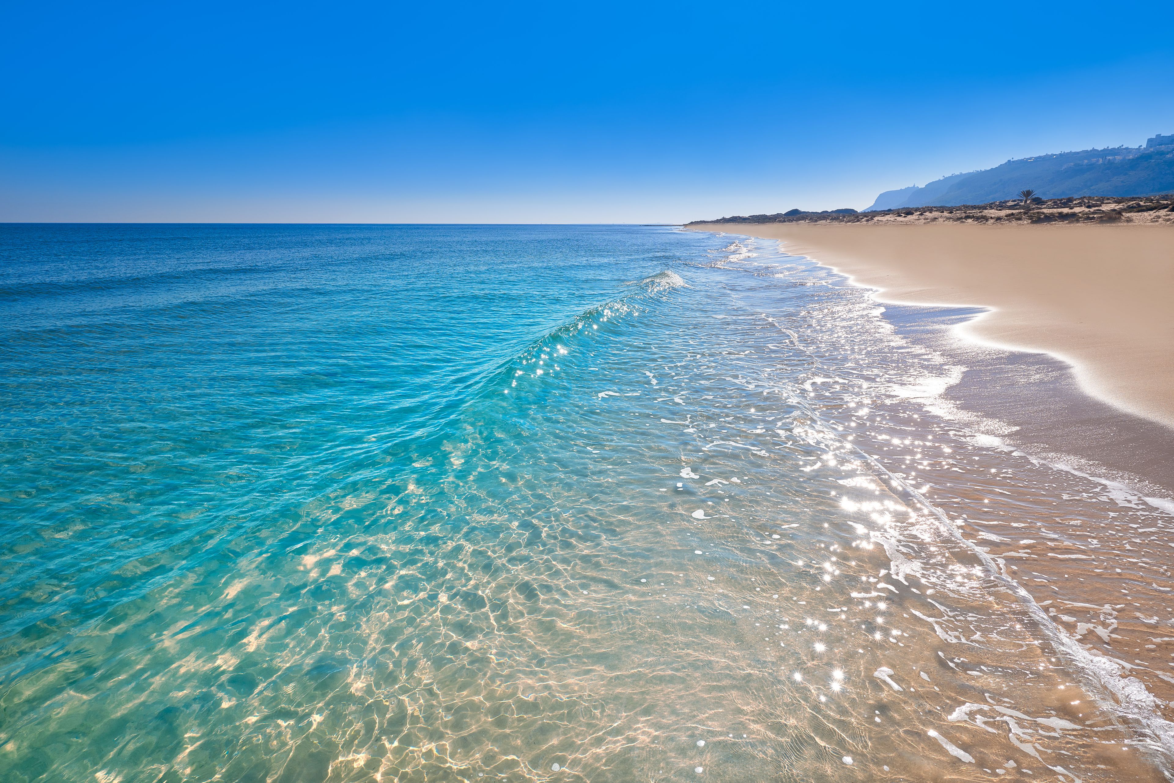Mar azul transparente con olas suaves, una playa arenosa y una cadena montañosa distante.