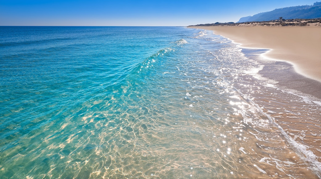 Mar azul transparente con olas suaves, una playa arenosa y una cadena montañosa distante.
