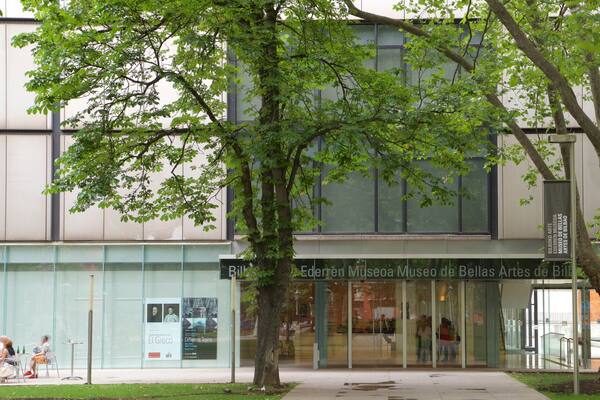 The exterior of the Ederren Museo de Bellas Artes de Bilbao, featuring a modern glass facade, a large tree in front, and a signboard with museum information.