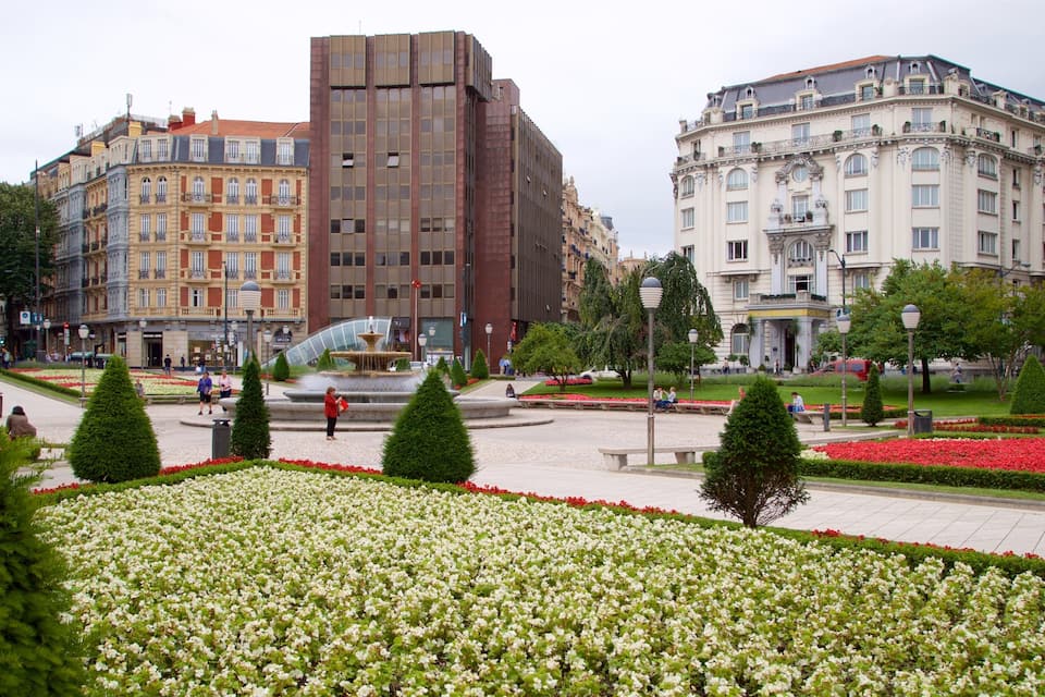 A city square with a fountain, flower beds, and historic buildings.