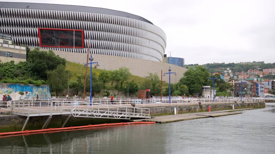 A waterfront promenade with a modern stadium, a bridge, and a river.