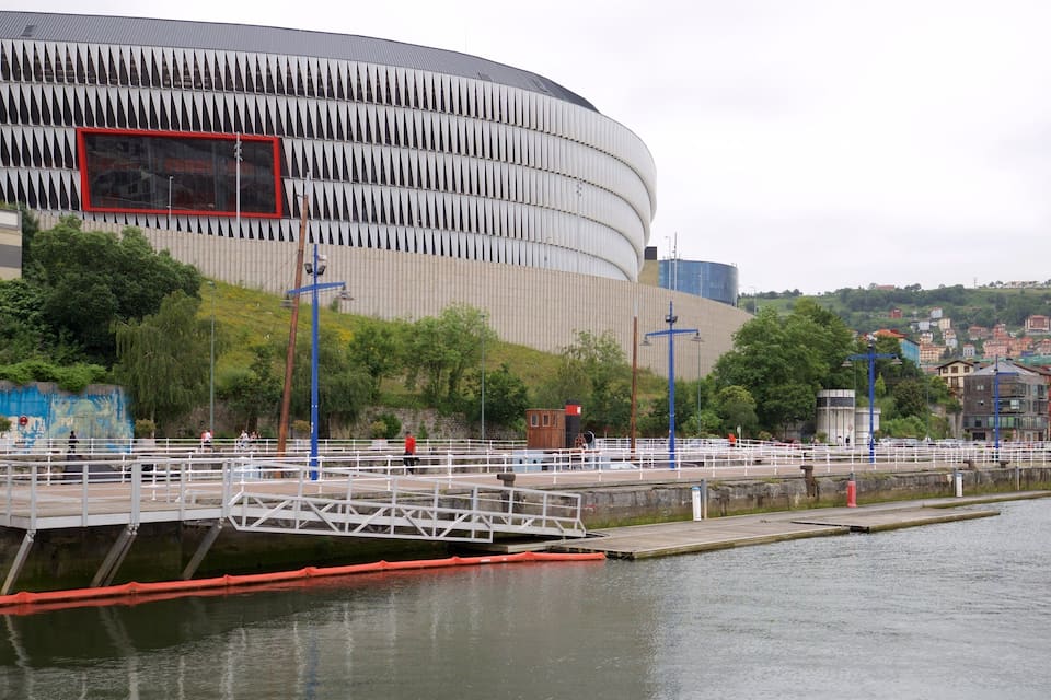 A waterfront promenade with a modern stadium, a bridge, and a river.