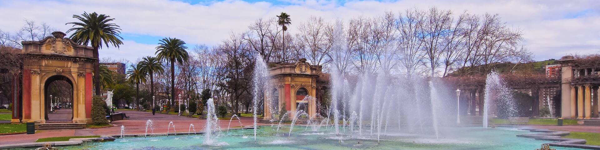 Una fuente con chorros de agua, rodeada por un césped cuidado y palmeras.