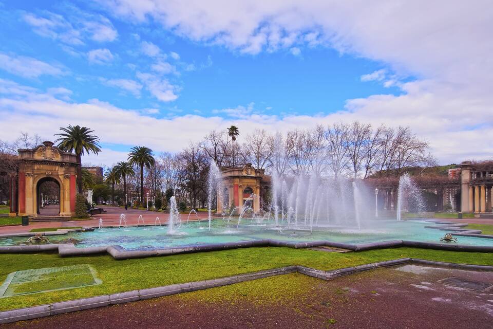Une fontaine avec des jets d’eau, entourée d’une pelouse bien entretenue et de palmiers.