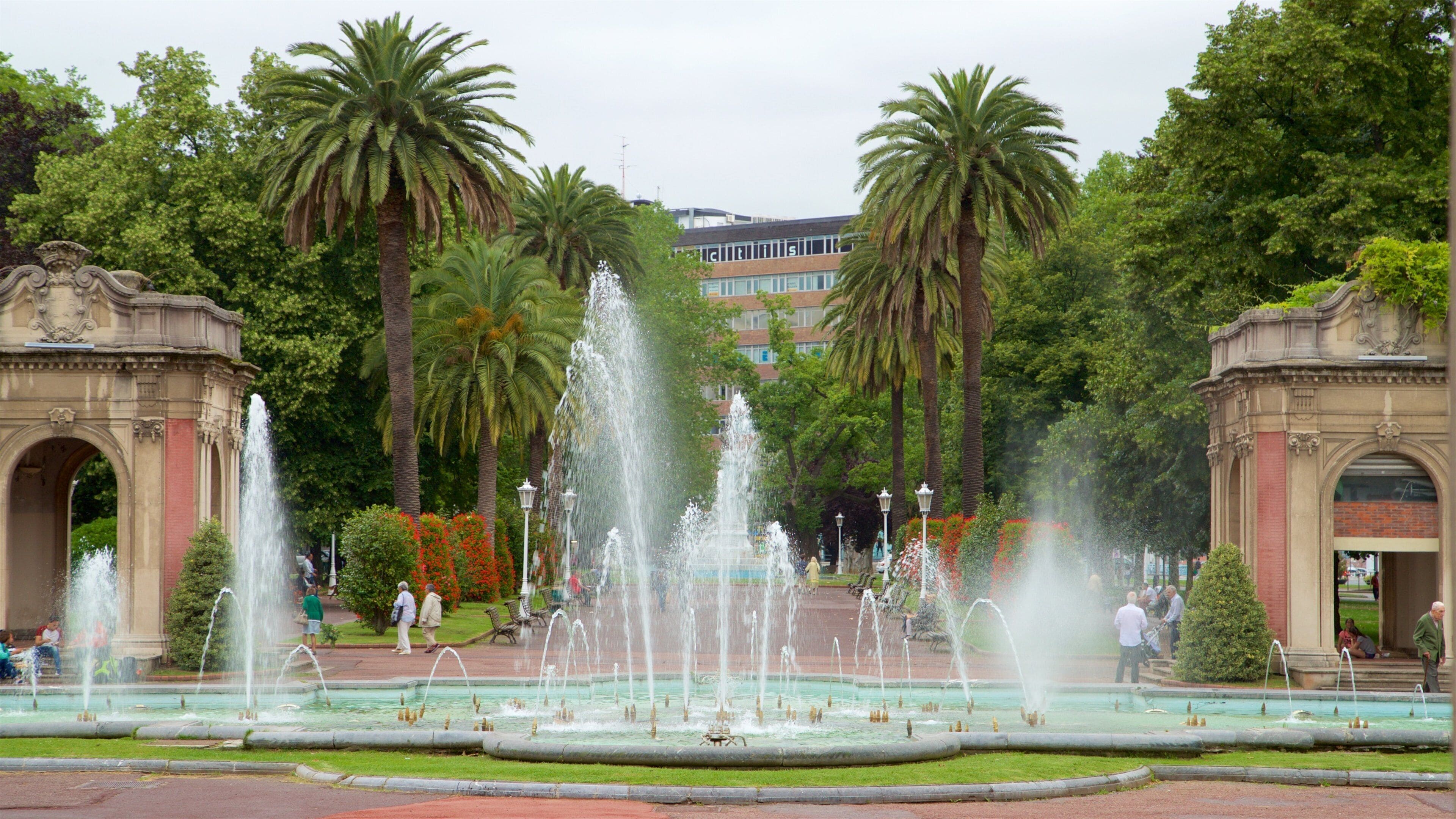 Una gran fuente con múltiples chorros de agua en un parque con palmeras y un edificio al fondo.