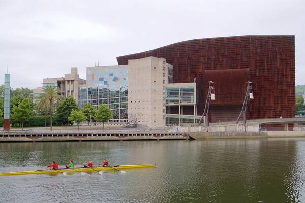 A modern building with a unique brick facade, a bridge, and a group of people rowing a yellow boat on a calm river.
