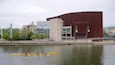 A modern building with a unique brick facade, a bridge, and a group of people rowing a yellow boat on a calm river.