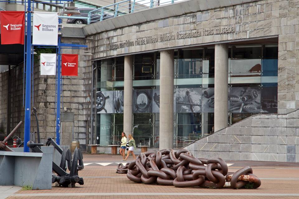Una gran escultura de cadenas de metal entrelazadas frente a un edificio con ventanas amplias y un letrero en un idioma extranjero.