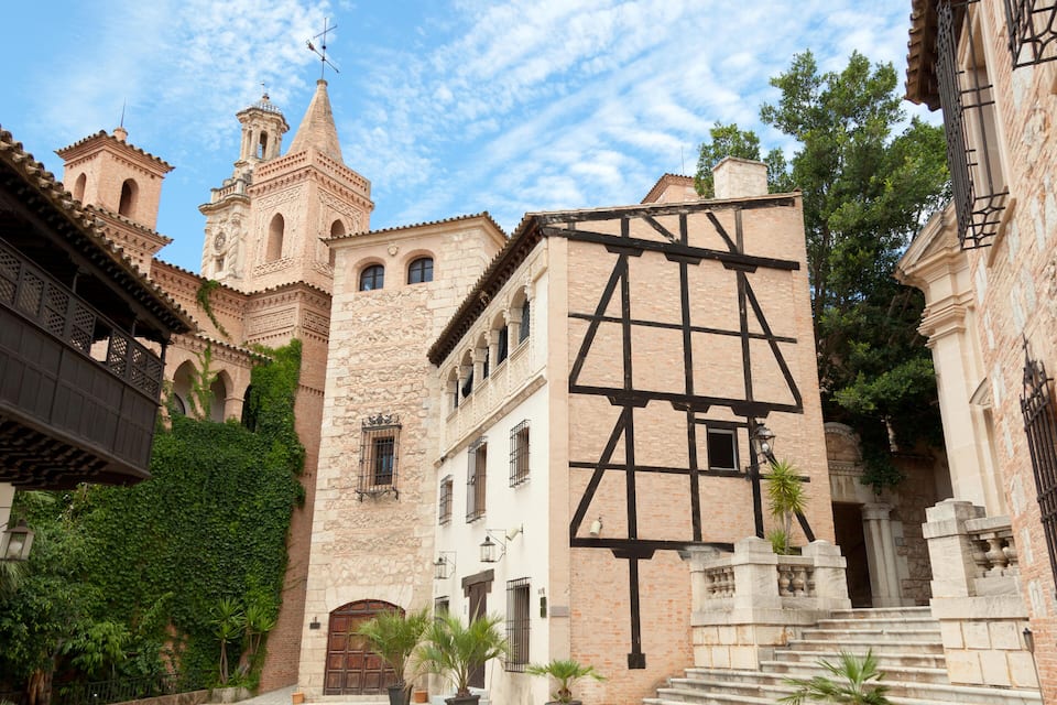 Un pati antic amb una torre campanar, un edifici amb estructura de fusta a la vista i plantes en testos.