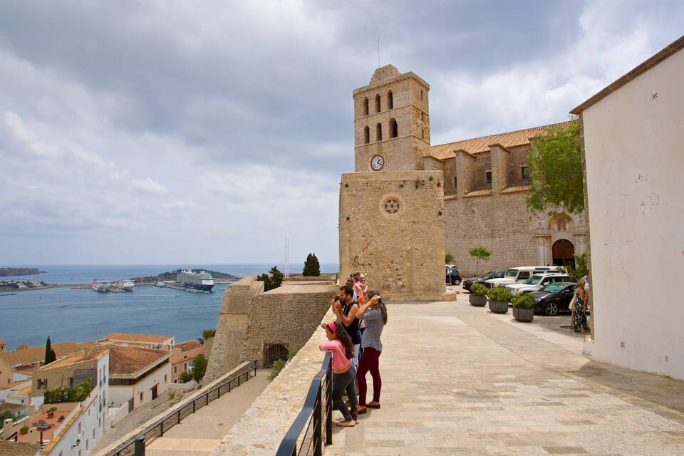 A coastal town with a prominent stone tower, a sea view, and parked cars along the promenade.
