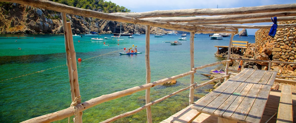 A wooden deck overlooking a turquoise sea with boats, a rocky coastline, and a clear sky.