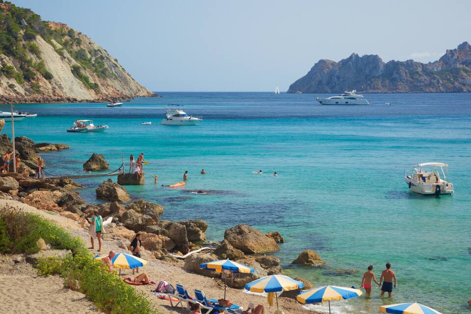 Een strand met helder turquoise water, een rotsachtige kustlijn en boten.