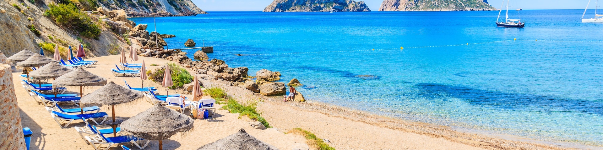 Una playa con sillas azules de descanso y sombrillas de paja, agua azul cristalina y acantilados rocosos.