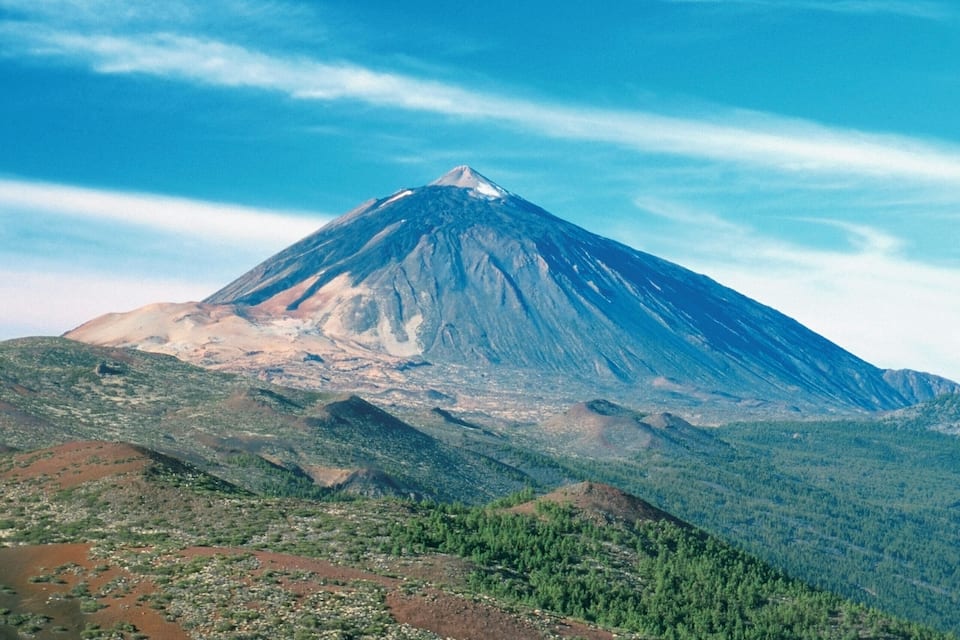Una montaña con cumbre nevada, rodeada de bosque verde y un río.