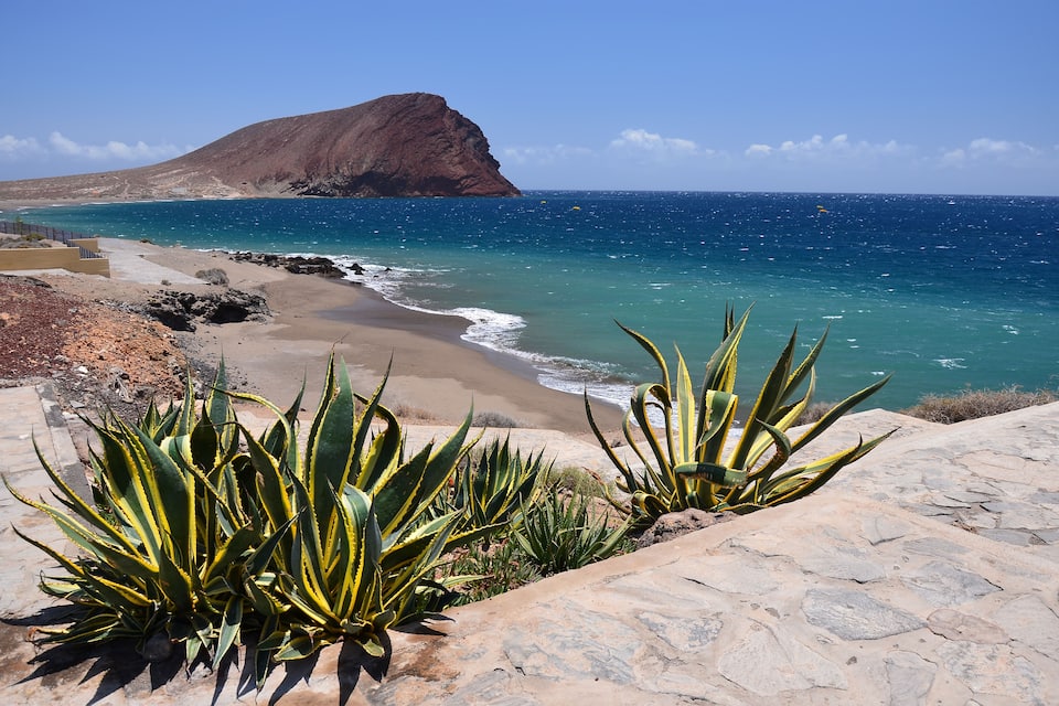 A coastal landscape with a rocky cliff, a sandy beach, and a clear blue sea.
