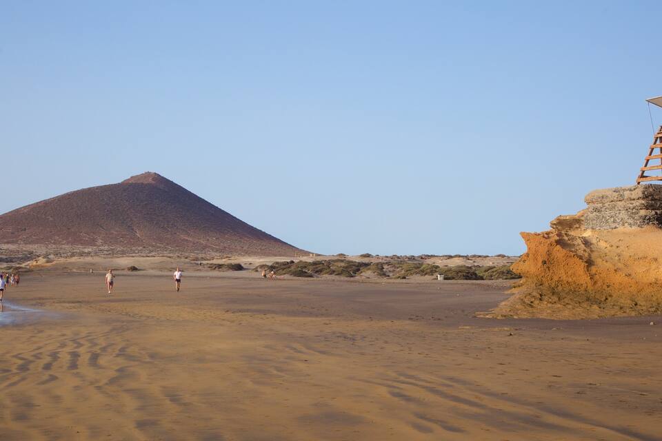 A desert landscape with a large hill, a wooden structure on a cliff, and people walking on the beach.