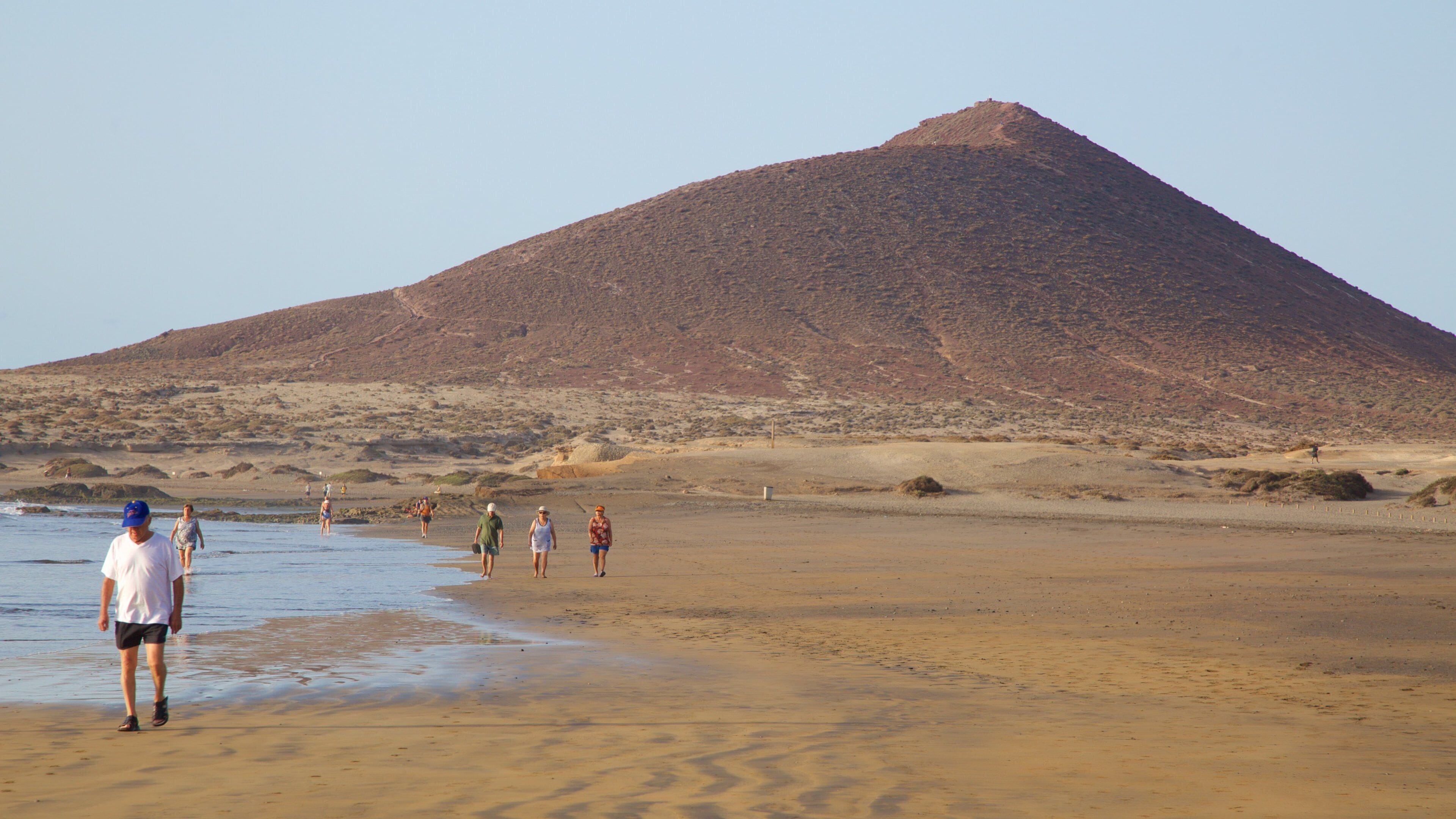 Uma praia com pessoas caminhando e uma grande colina ao fundo.